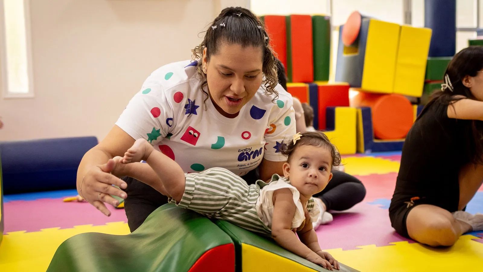 Professora apoia um bebê que se equilibra em uma rampa macia durante atividade na Baby Gym Manaus.