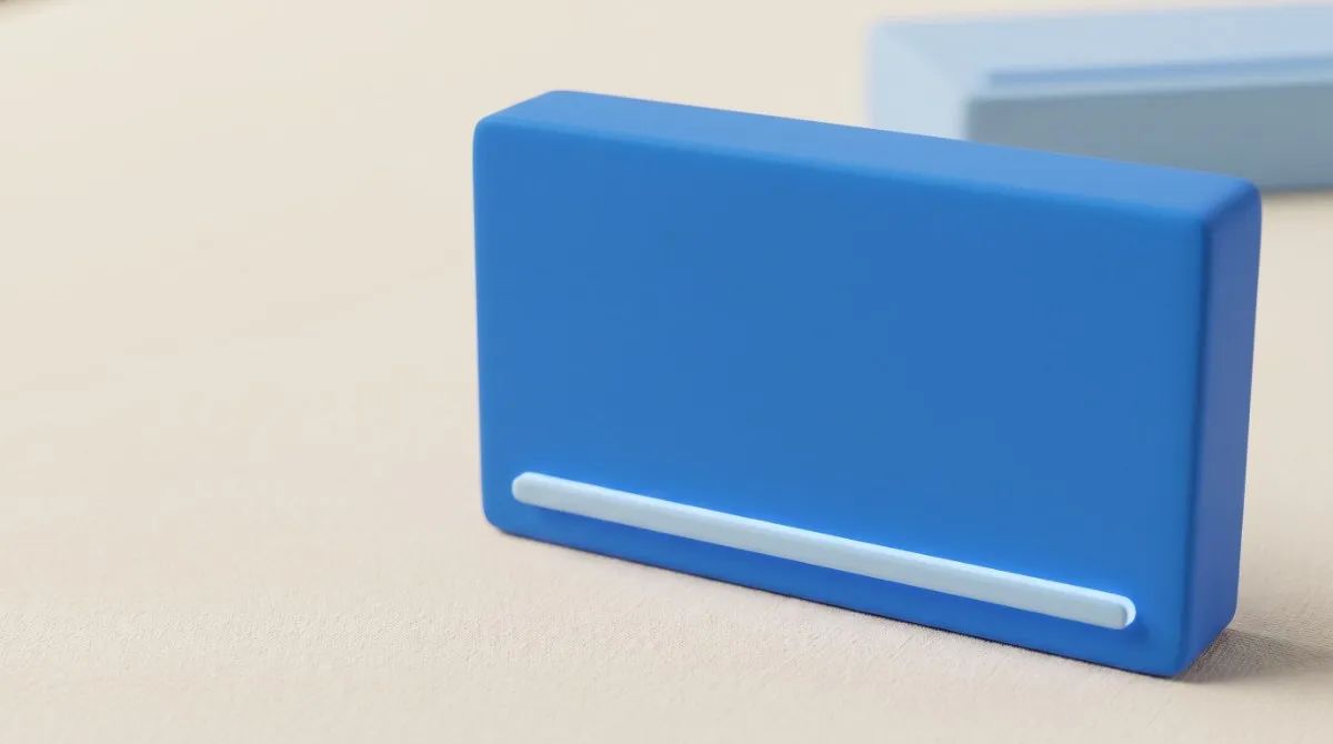 Close de uma caixa de remédio azul sobre uma mesa creme, sem rótulo, com uma única linha clara em relevo perto da base, onde ficaria o nome da molécula; outra caixa, azul-clara, fora de foco atrás.